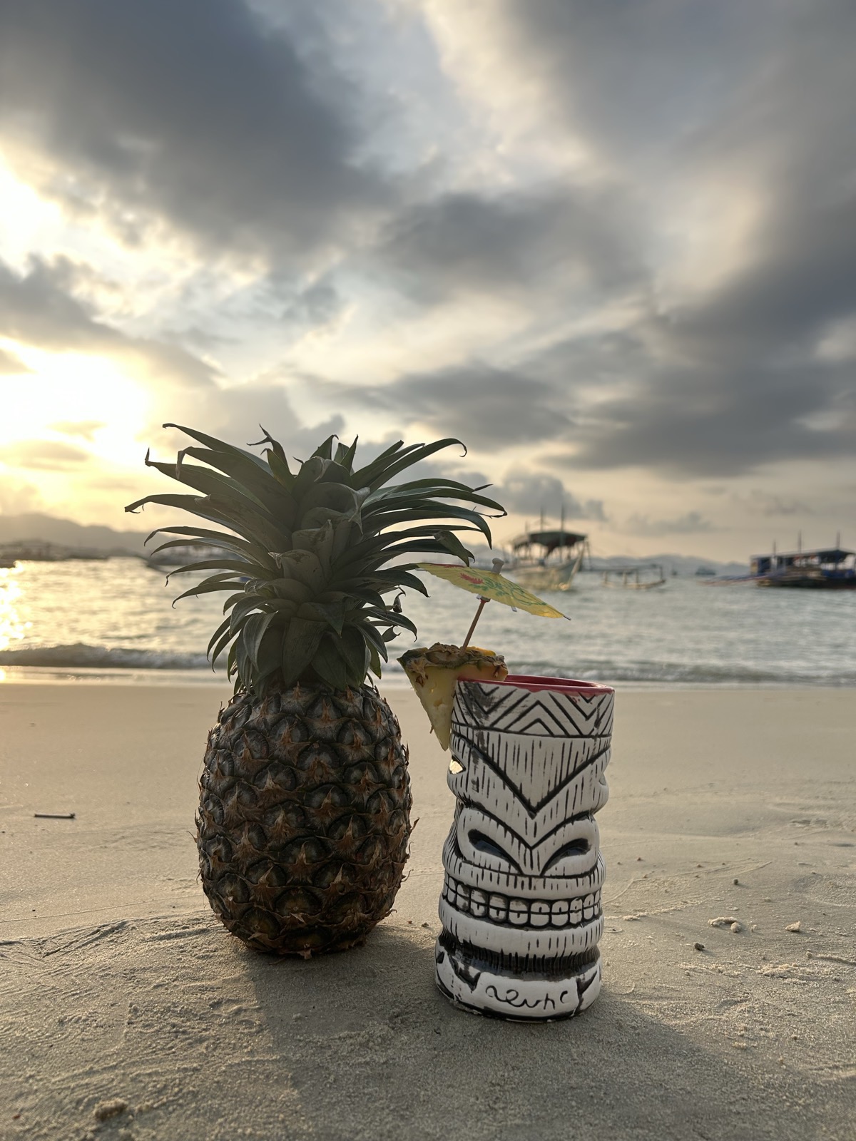 Signature carved tiki mug and whole-pineapple cocktail on the sand at sunset, Mai Tai Port Barton